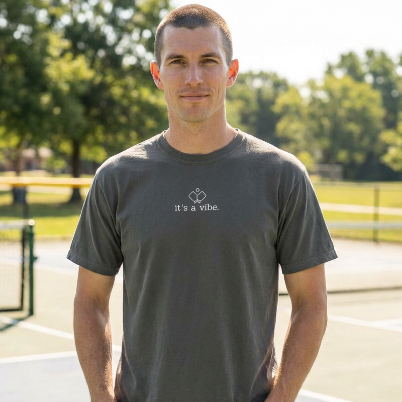 Man wearing a gray comfort color pickleball t-shirt that says it's a vibe standing outdoors on a sunny day.