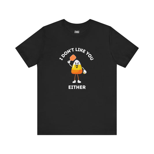 A black short-sleeve t-shirt with a graphic of a cartoon candy corn character holding a pickleball paddle and a pickleball and the text 'I DON'T LIKE YOU EITHER' printed on the front, displayed against a solid white background.