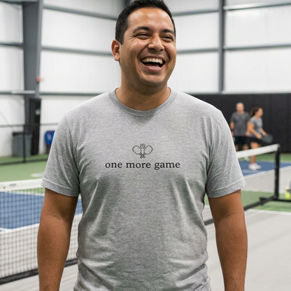 A man laughing on an indoor pickleball court wearing a heather gray t-shirt with crossed paddles and the text 'one more game'.