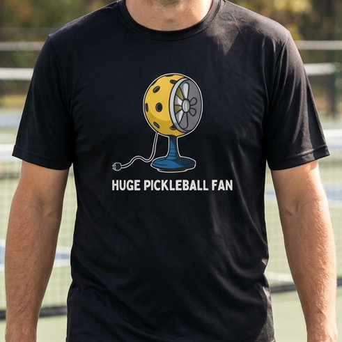 A man standing on a blue pickleball court wearing a black t-shirt with a yellow and blue pickleball graphic and the text 'HUGE PICKLEBALL FAN'.