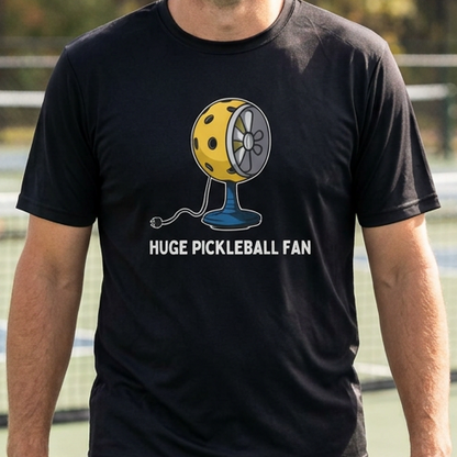 A man standing on a blue pickleball court wearing a black t-shirt with a yellow and blue pickleball graphic and the text 'HUGE PICKLEBALL FAN'.