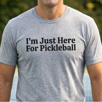 A man standing on an outdoor pickleball court wearing a heather gray t-shirt with the black printed text 'I'm Just Here For Pickleball'.