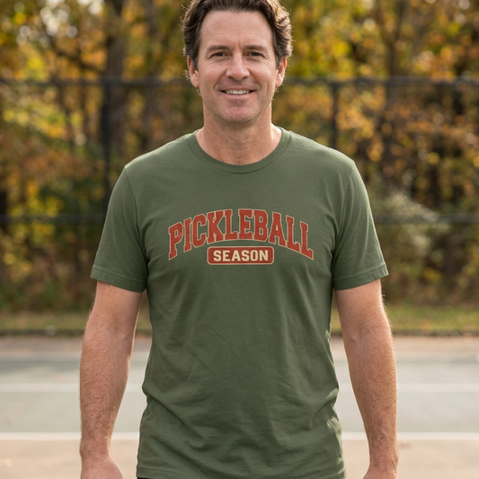 A man standing on an outdoor pickleball court wearing an olive green picklebal lt-shirt with 'PICKLEBALL SEASON' 