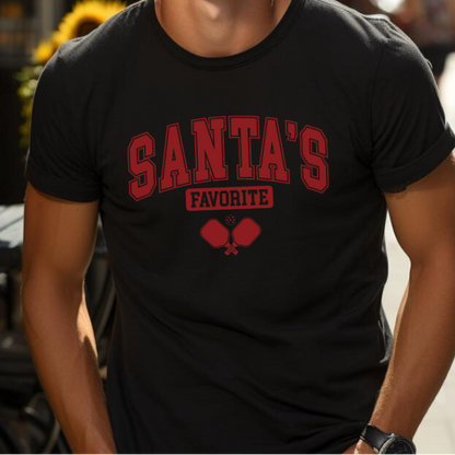 A man wearing a black short-sleeve pickleball t-shirt with 'SANTA'S FAVORITE' printed in red collegiate-style lettering and a red pickleball paddle graphic below, shown in a lifestyle setting.