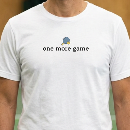 A person wearing a white short-sleeve crewneck t-shirt  featuring a small graphic of a pickleball paddle and pickleball above the black text 'one more game' on the chest.