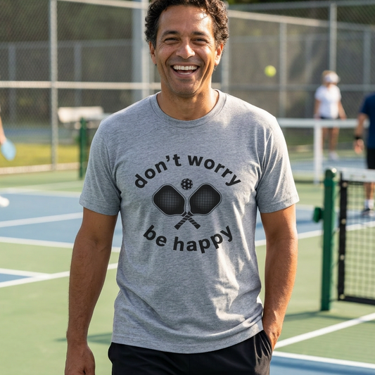 A smiling man on a pickleball court wearing a heather gray t-shirt with a black graphic that reads 'don't worry be happy' around crossed pickleball paddles.  