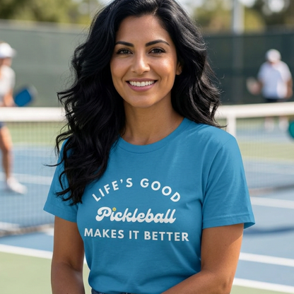 A woman sitting on a bench at a pickleball court wearing a blue graphic t-shirt that reads 'LIFE'S GOOD Pickleball MAKES IT BETTER' in white text.