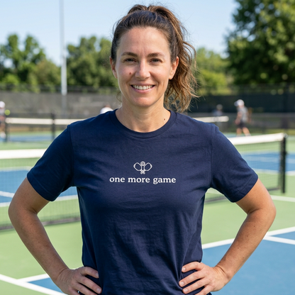 A woman standing on a pickleball court wearing a navy blue t-shirt with a crossed pickleball paddles graphic and the text 'one more game'.