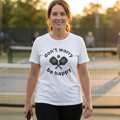A woman standing on a pickleball court wearing a white pickleball t-shirt with the text 'don't worry be happy' and crossed pickleball paddles