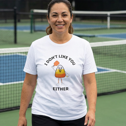 A woman standing on an outdoor pickleball court wearing a white graphic t-shirt with a cartoon pickleball candy corn character and the text 'I DON'T LIKE YOU EITHER'.