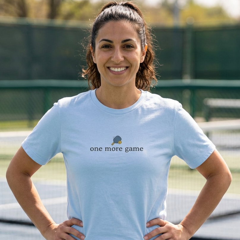 A woman stands on a pickleball court wearing a light blue crewneck pickleball t-shirt with the text 'one more game' and a small pickleball graphic printed above it.