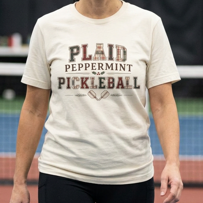 A woman stands on an indoor pickleball court wearing a cream-colored graphic t-shirt that reads 'PLAID PEPPERMINT PICKLEBALL' in dark lettering.