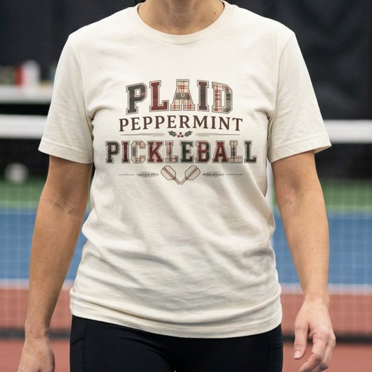 A woman stands on an indoor pickleball court wearing a cream-colored graphic t-shirt that reads 'PLAID PEPPERMINT PICKLEBALL' in dark lettering.