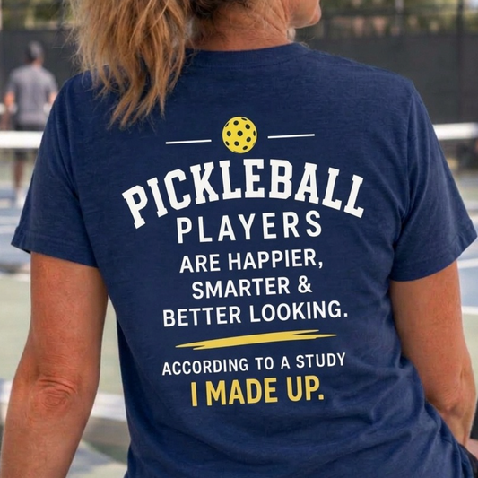 A woman viewed from behind wearing a navy blue pickleball t-shirt with a humorous pickleball graphic and text on the back, set against a blurred outdoor pickleball court background.