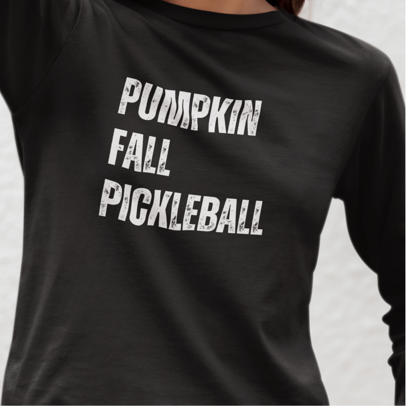 A woman wearing a black long-sleeve pickleball t-shirt with the words 'PUMPKIN FALL PICKLEBALL' printed in distressed white block letters on the front, photographed against a light textured wall.
