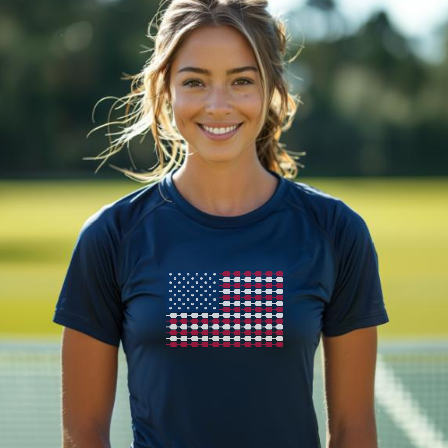 A woman wearing a navy performance t-shirt with a graphic of the American flag designed with pickleballs and paddles on it, standing on a pickleball court.