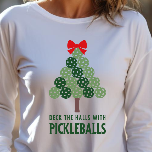 A woman with blonde hair smiling outdoors, wearing a white long-sleeve shirt with a green pickleball Christmas tree graphic and the text 'DECK THE HALLS WITH PICKLEBALLS'.