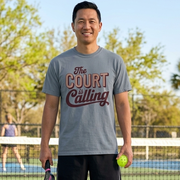 Man on a pickleball court holding a paddle and pickleball, wearing a 'The Court is Calling' t-shirt.