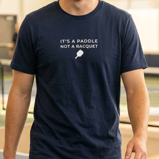 Man wearing a navy blue pickleball t-shirt that says it's a paddle not a racquet an indoor pickleball court