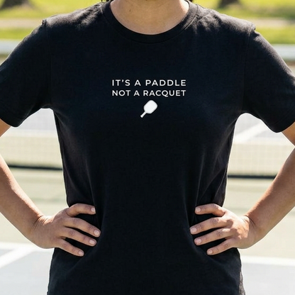 Woman wearing a black pickleball t-shirt on a pickleball court that says it's a paddle not a racquet