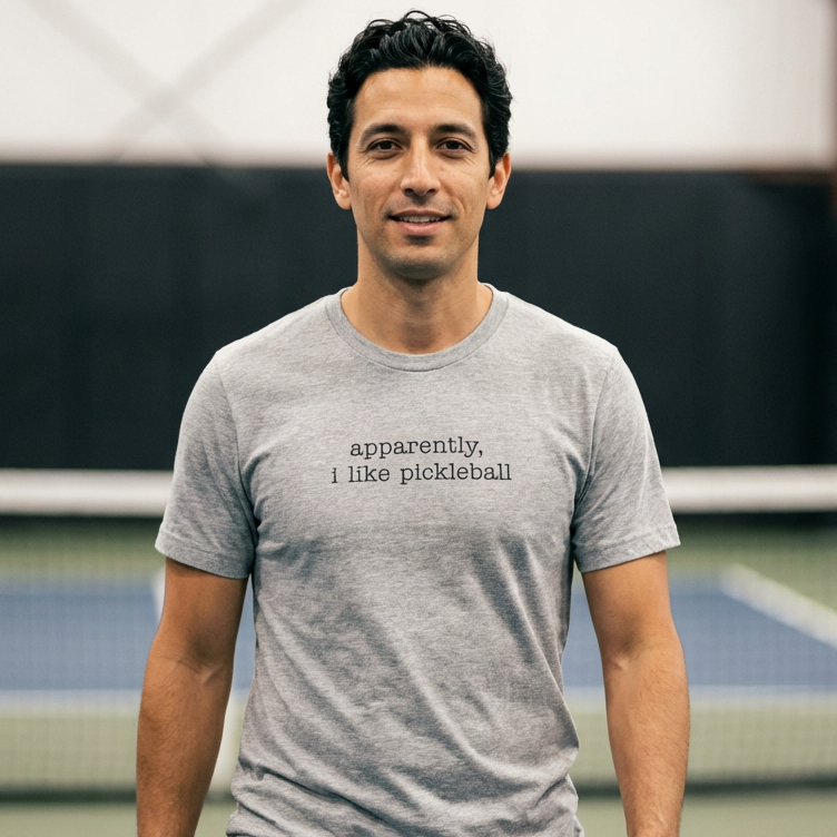 Man wearing a gray t-shirt with that says apparenetly I like pickleball on a pickleball court