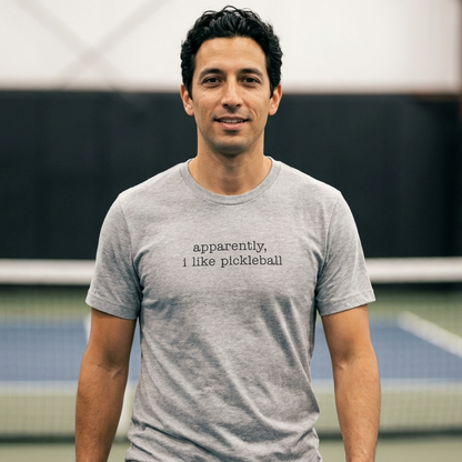 Man wearing a gray t-shirt with that says apparenetly I like pickleball on a pickleball court